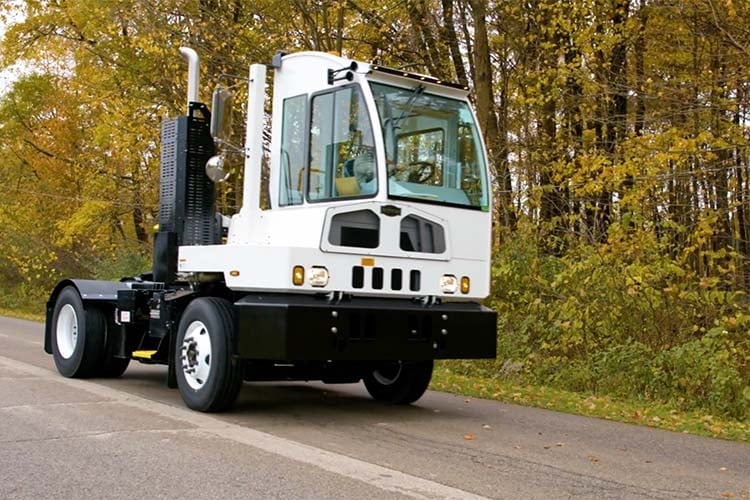 White and black terminal tractor with lift gate equipment parked on road with autumn trees in background